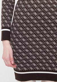 Primo piano di un vestito a maglia marrone con un motivo a diamante bianco ripetuto e logo, che mostra la mano sinistra e le maniche a coste con strisce bianche.