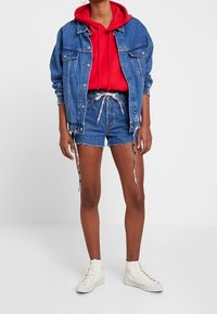 Denim jacket in blue, red hooded sweatshirt underneath, and blue shorts with drawstrings. White sneakers complete the outfit.