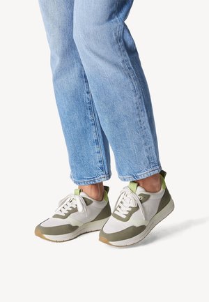 Personne portant un jean bleu clair et des baskets blanches avec des accents verts, debout sur un fond blanc.