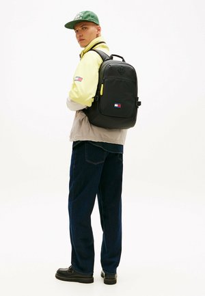 Joven con gorra verde, chaqueta amarilla y beige, vaqueros oscuros, zapatos negros y mochila negra con un pequeño parche de logotipo.