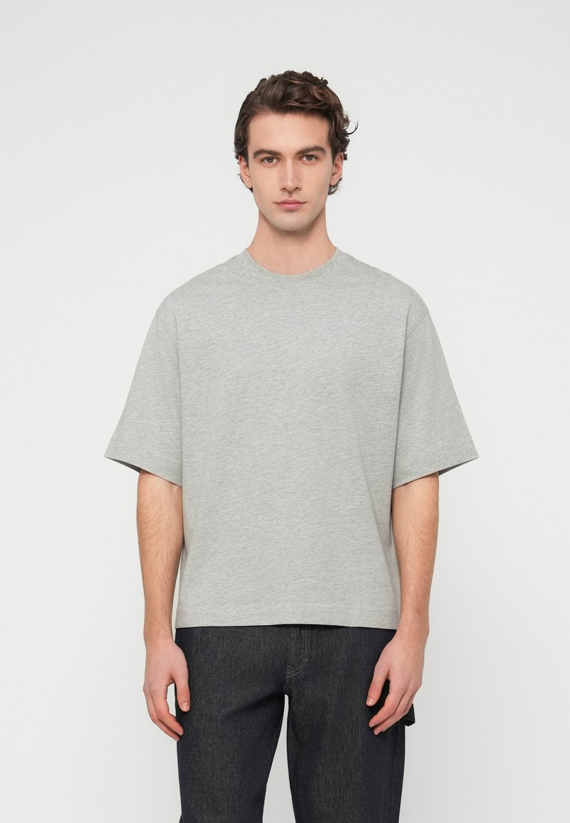 Γκρι βαμβακερό μπλουζάκι με κοντά μανίκια, στρογγυλή λαιμόκοψη και άνετη εφαρμογή. Το ύφασμα φαίνεται λείο και χωρίς ιδιαίτερα χαρακτηριστικά, χωρίς ορατά μοτίβα ή διακοσμήσεις.