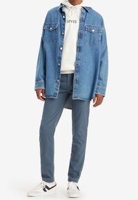 Denimskjorta i ljusblått med två bröstfickor och knappstängning, bärs över en vit hoodie med "LEVI'S" i grön text, tillsammans med grå byxor och sneakers.