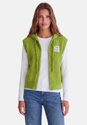 Femme portant un gilet à capuche zippé duveteux vert vif sur une chemise blanche à manches longues et un jean bleu, regardant vers l'avant avec une expression neutre.