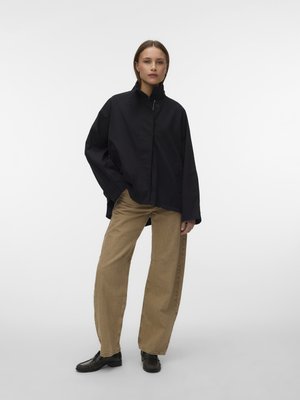 Naine seisab, kandes lahtist musta nööpidega jopet, beeži laiade pükstega ja mustade kingadega üksluisel heledal taustal.