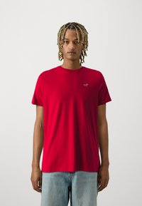 Rotes Baumwoll-T-Shirt mit rundem Hals und kurzen Ärmeln; hat ein kleines Logo auf der linken Brust; getragen mit hellblauen Jeansshorts.