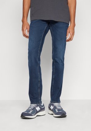 Personne portant un jean slim bleu et des baskets New Balance grises et bleu marine, debout devant un fond clair uni.