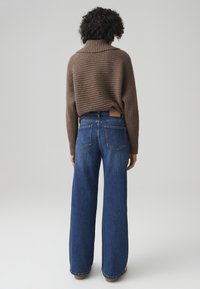 Maglione a coste marrone con colletto alto, abbinato a jeans in denim blu a gamba larga con tasche posteriori e dettagli di cucitura discreti.