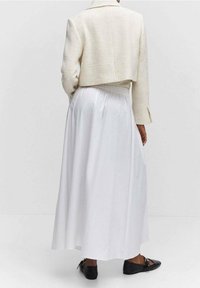 Valkoinen maxi-hame, jossa on virtaava rakenne, yhdistettynä lyhytlahkeiseen, vaaleanväriseen tweed-takkiin, jossa on kaulus ja pitkät hihat. Mustat matalat kengät.