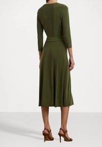 Kvinna klädd i en olivgrön, långärmad midi-klänning med en figurnära midja och bruna högklackade sandaler, stående mot en vit bakgrund.