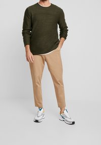 Pull texturé vert olive à col rond, associé à un pantalon beige fuselé et des baskets blanches avec des accents bleus, jaunes et noirs.