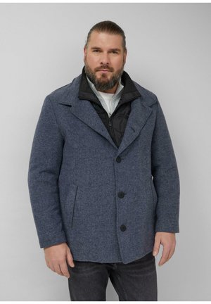 Mann mit Bart, der einen blauen, geknöpften Kurzmantel über einer schwarzen Reißverschlussjacke und einem weißen Hemd trägt, steht vor einem schlichten hellgrauen Hintergrund.