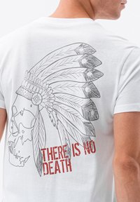 Vit t-shirt med en detaljerad grafik av ett skelett som bär en amerikansk urinvånartäckning, med texten "DET FINNS INGEN DÖD" i röda bokstäver.