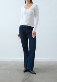 Vit ribbad långärmad topp med knappraderad accent, kombinerad med mörkblå vida jeans, bärs tillsammans med svarta klackade stövlar.