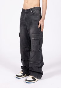 Mężczyzna noszący luźne czarne spodnie cargo z wieloma kieszeniami oraz czarno-białe trampki, stojący na białym tle.