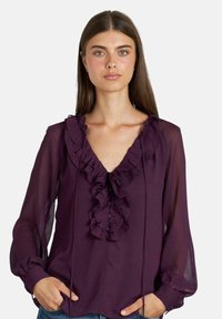 Blusa viola con accentuazione a volant sul collo, maniche lunghe e trasparenti, e dettagli con bottoni. Tessuto morbido con una sottile texture.
