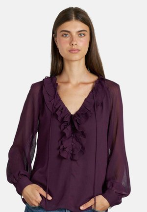 Blouse violette avec un accent volé au niveau du col, manches longues transparentes et détails en boutons. Tissu doux avec une texture subtile.