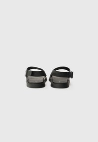 Svarte sandaler med et elegant design som har en bred stropp og justerbar spenne. Mykt, strukturert fotbed og minimalistisk gummisåle.