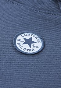 O țesătură gri cu o pată albă circulară care prezintă o stea neagră și textul "CONVERSE ALL STAR". Textură netedă cu margini cusute.