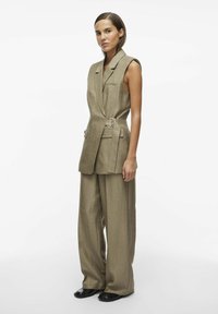 Chaleco beige ajustado con cuello, bolsillos laterales y cintura ceñida con cinturón, combinado con pantalones beige de corte holgado y zapatos negros.