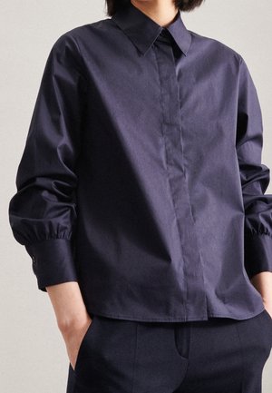 Person iført en marineblå skjorte med lange ærmer og knapper, med manchet detaljer, hænderne i lommerne, mod en ensfarvet baggrund.