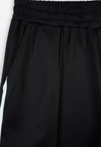 Czarne dresowe spodnie z elastycznym pasem, bocznymi kieszeniami i białymi akcentującymi paskami po bokach. Miękka, gładka tekstura.