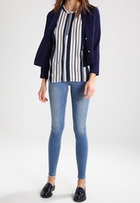 Blazer bleu marine porté sur un chemisier blanc à rayures bleu marine, associé à un jean bleu ajusté et des mocassins bleu marine. Tissu lisse, design sur mesure.