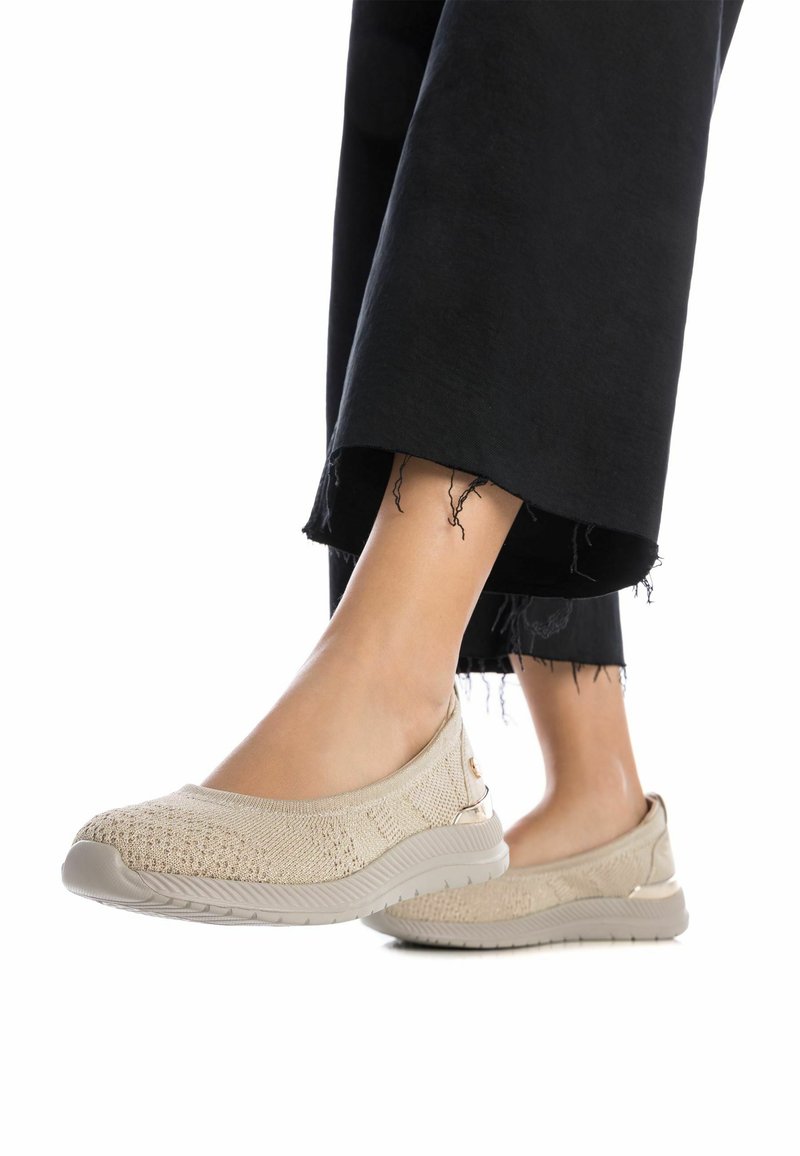 Zapatos slip-on beige con parte superior de punto texturizado, suela flexible y un sutil acento metálico. Combinados con pantalones negros de dobladillo deshilachado.
