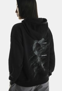 Mustvalge kapuutsiga pusa, millel on tekstuuriga tagaküljel valge graafika, mis sisaldab punktmustreid ja tekstilisi detaile, sealhulgas "ON RUNNING" ja "SWISS ENGINEERING."