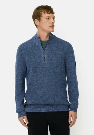 Ung mand iført en blå strikket quarter-zip sweater over en mørk skjorte, stående og kigger til siden mod en ensfarvet baggrund.
