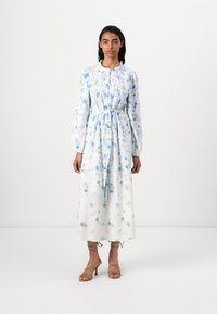 O vestido floral branco apresenta flores azuis e verdes, mangas longas, uma cintura atada e um colarinho repolgado. Complementado com sandálias de tiras em tom bege.