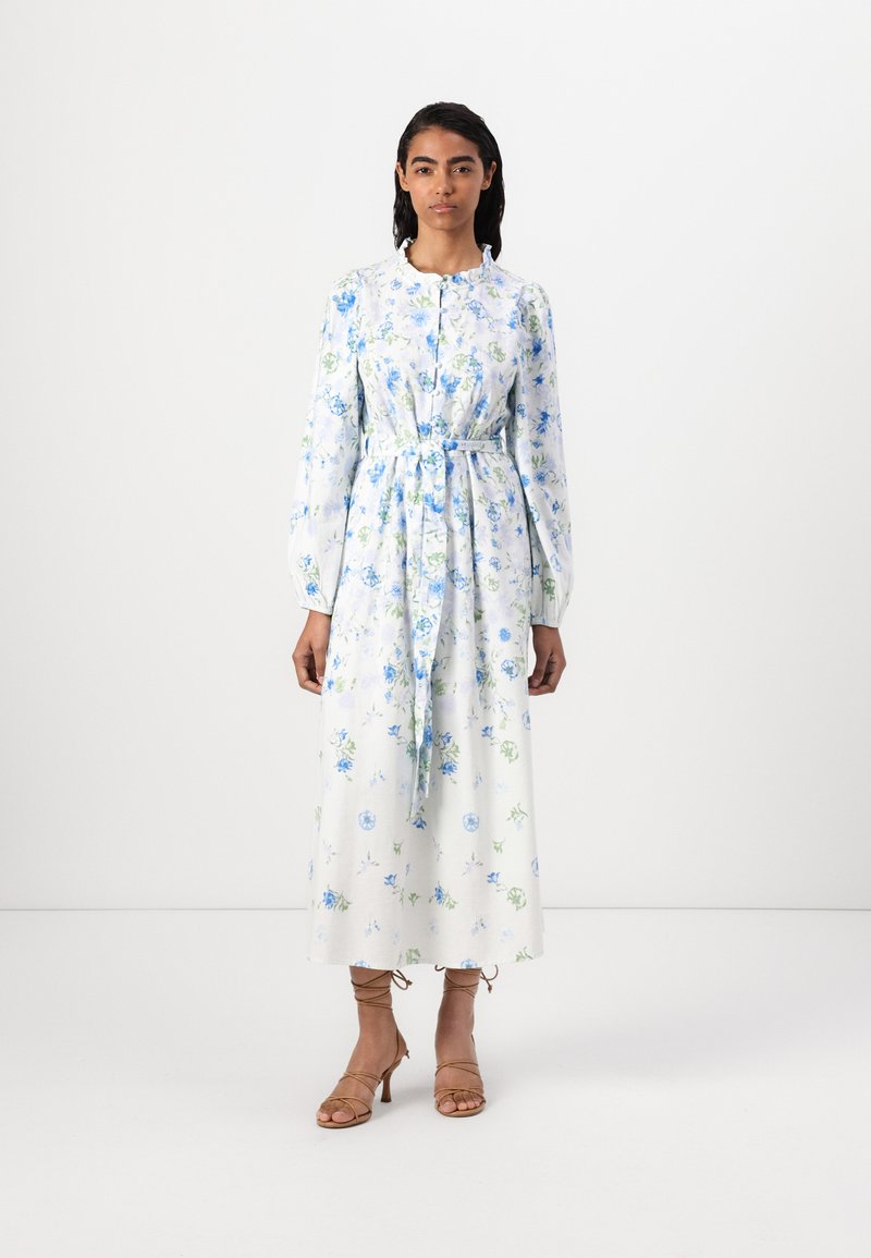 O vestido floral branco apresenta flores azuis e verdes, mangas longas, uma cintura atada e um colarinho repolgado. Complementado com sandálias de tiras em tom bege.