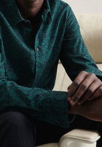 Camicia a maniche lunghe di colore verde scuro con un motivo floreale, realizzata in tessuto testurizzato, arricchita da bottoni neri, indossata con pantaloni scuri.