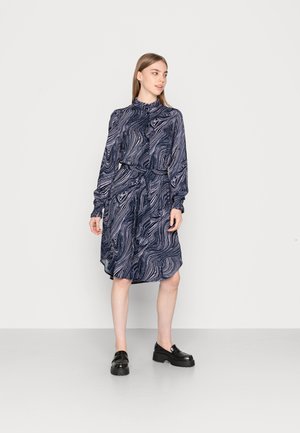 Vestido azul-marinho com padrões brancos em espiral, decote alto, mangas longas e cintura com cinto. Sapatos de plataforma pretos complementam o conjunto.