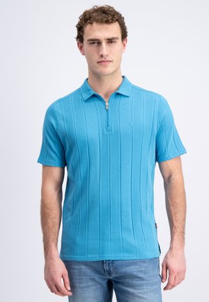Uomo con polo lavorata a maglia azzurra chiara a maniche corte con zip e jeans blu, in piedi davanti a uno sfondo bianco uniforme.