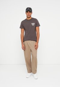 Brązowa koszulka z krótkim rękawem z białym nadrukiem, beżowe spodnie szerokie i białe sneakersy. Czarna czapka z logo podkreślająca stylizację.