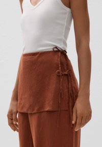 Linen top v rustovej farbe s výstrihom do V a detailom na viazanie na boku. Látka vyzerá jemne a textúrovane, má voľný strih a vrstvenie na leme.