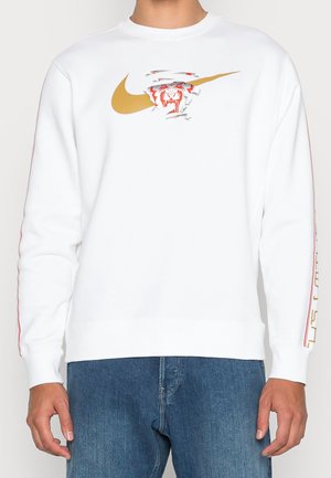 Biała bluza z długim rękawem z złotym logo Nike i czerwonym logo w kształcie twarzy tygrysa na piersi, noszona z niebieskimi dżinsami.