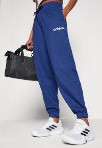 Modré bavlněné tepláky s elastickými lemy a páskem, s bílým logem Adidas. Kombinovány s bílými teniskami a černou sportovní taškou.