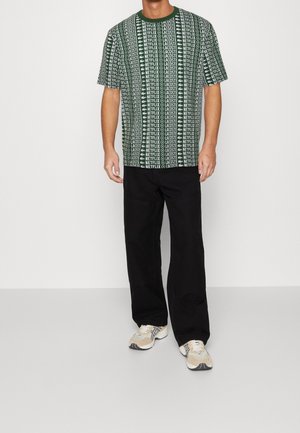 Grüne T-Shirt mit Kurzarm, das ein sich wiederholendes Textmuster hat. Kombiniert mit legere schwarze Hose und beigen Turnschuhen.