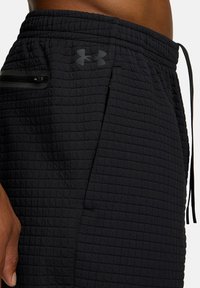 Svarta quiltade joggers med en texturerad tyg, elastisk midja, sidofickor med dragkedja och en subtil logotyp nära midjan.