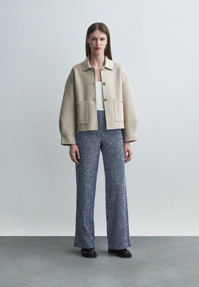 Γυναίκα που στέκεται φορώντας μπεζ σακάκι με κουμπιά, λευκό τοπ, μπλε φαρδιά υφασμάτινη παντελόνια με υφή και σκούρα παπούτσια σε απλό γκρι φόντο.