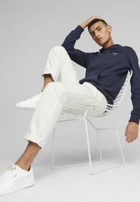 Marineblaues Sweatshirt, weiße Hose, weiße Sneaker. Modell sitzt in einem weißen Drahtstuhl, vor einem schlichten, modernen Hintergrund.