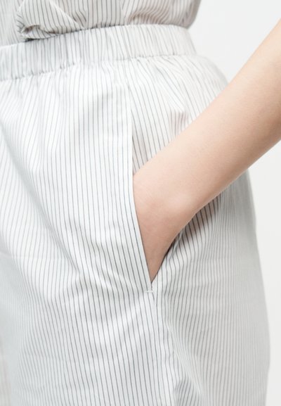 Χέρι που ακουμπά μέσα στην τσέπη άσπρου παντελονιού με λεπτές κάθετες μαύρες ρίγες και ελαστική ζώνη μέσης.