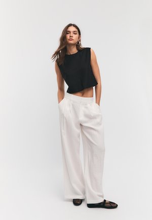 Kobieta stojąca z rękami w kieszeniach, ubrana w czarny, bezrękawnikowy top odsłaniający brzuch, szerokie białe spodnie i czarne siateczkowe baleriny na białym tle.