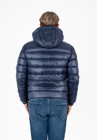Marineblauwe gewatteerde jas met een capuchon, met gekwikste delen, een glanzende, gladde textuur en een getailleerde pasvorm. Draagt met blauwe jeans.
