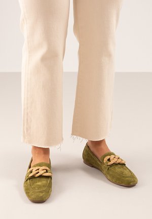 Mocasines de ante verde con detalles trenzados en beige. Punta redondeada y suela plana. Llevados con pantalones color crema, ligeramente deshilachados en el dobladillo.