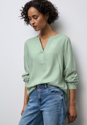 Blusa verde menta con scollo a V, maniche lunghe raccolte sui polsi, abbinata a jeans in denim azzurro chiaro. Tessuto liscio, vestibilità comoda.