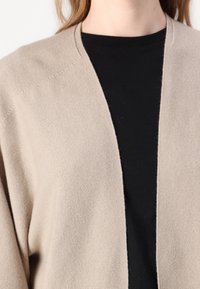 Beige cardigan med løs, åpen frontdesign, laget av strikket stoff. Den har en glatt tekstur og raglanermer, lagd over en svart topp.