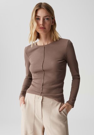Bruine langemouwen top met een textuur van ribbels, met een centrale naad en een ronde halslijn. Gecombineerd met lichtbeige trousers.