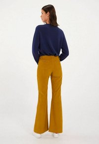 Pantalons en velours côtelé jaune évasés avec poches arrière et passants de ceinture, associés à une chemise à manches longues bleu marine. Des baskets blanches complètent le look.